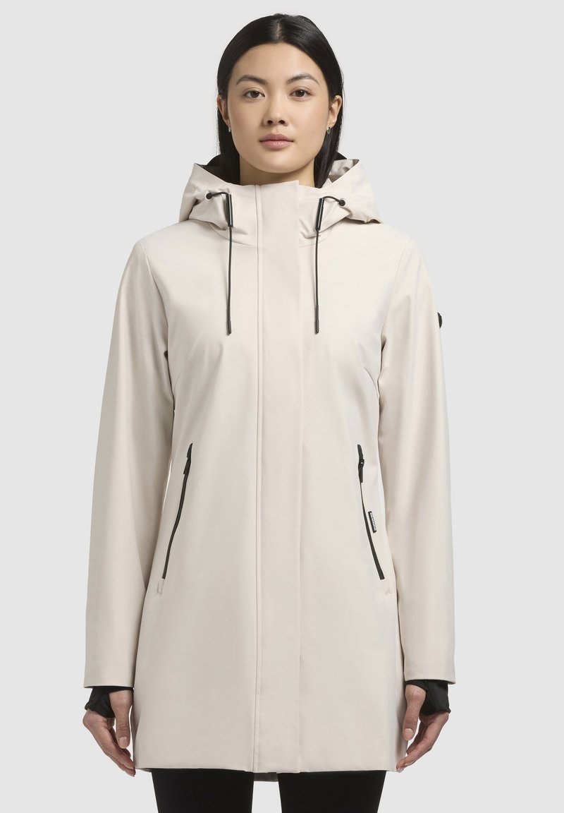 Manteau imperméable beige clair avec capuche, doté de poches zippées noires, d'un design épuré et de manches longues avec des poignets intérieurs pour plus de chaleur.