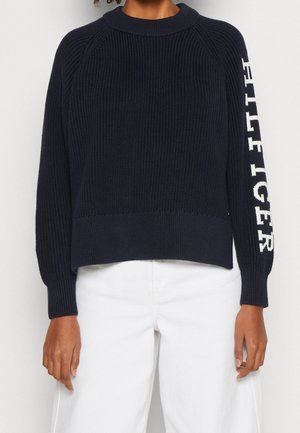 Zwarte gebreide trui met witte "HILFIGER" tekst op de linker mouw, gedragen met hooggetailleerde witte broek, handen ontspannen langs het lichaam.