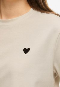 Beżowa koszula z krótkim rękawem wykonana z miękkiego materiału, z czarnym haftem w kształcie serca po lewej stronie klatki piersiowej.