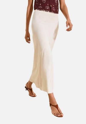 Person går iført en kremfarvet lang nederdel, brune åbne hælsandaler og en cropped mørkerød vest med knappedetaljer.