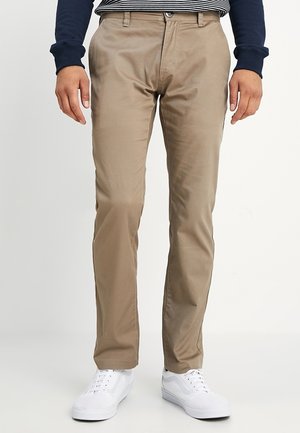 Uomo con chinos beige a gamba dritta, sneaker bianche e una camicia a maniche lunghe blu navy con orlo a righe visibile.