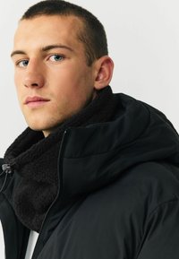 Čierna vypchávkovaná bunda s veľkou kapucňou a chlpatým čiernym golierom. Tkanina sa zdá byť hladká, s predným zipsom a nastaviteľnou šnúrkou na krku.