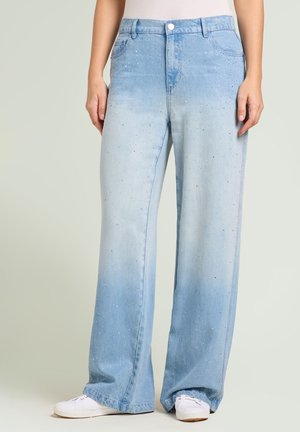 Donna che indossa jeans a gamba larga azzurro chiaro con piccole applicazioni di strass e sneakers bianche, in piedi contro uno sfondo semplice.