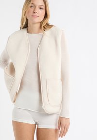Fleecová vesta v světlé krémové barvě s zaoblenými okraji, kontrastním lemováním a dvěma předními kapsami, vrstvená přes tenké tričko s dlouhými rukávy.