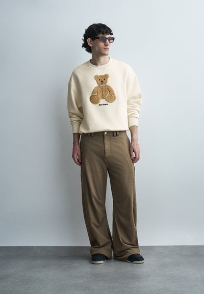 Άνδρας με μπεζ φούτερ με γραφικό αρκουδάκι, καφέ παντελόνια φαρδιάς γραμμής, μαύρα παπούτσια και γυαλιά ηλίου, στέκεται μπροστά σε απλό γκρι τοίχο.