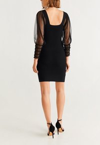 Schwarzes, enges Kleid mit transparenten Puffärmeln, einem quadratischen Rückenausschnitt und strukturierten Ärmelakzenten. Kombiniert mit schwarzen High-Heels.