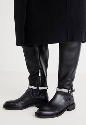Bottes en cuir noires à hauteur de genou avec fermetures éclair latérales et brides cheville ornées de strass, portées avec un manteau noir.