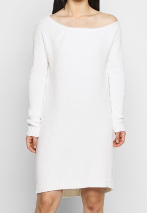 Femme portant une longue robe-pull en maille côtelée blanche avec un large décolleté bardot et des manches longues.