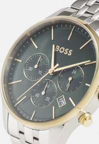 Reloj de acero inoxidable plateado con una esfera verde oscura, acentos dorados, tres subesferas y una pantalla de fecha a las 6 en punto.