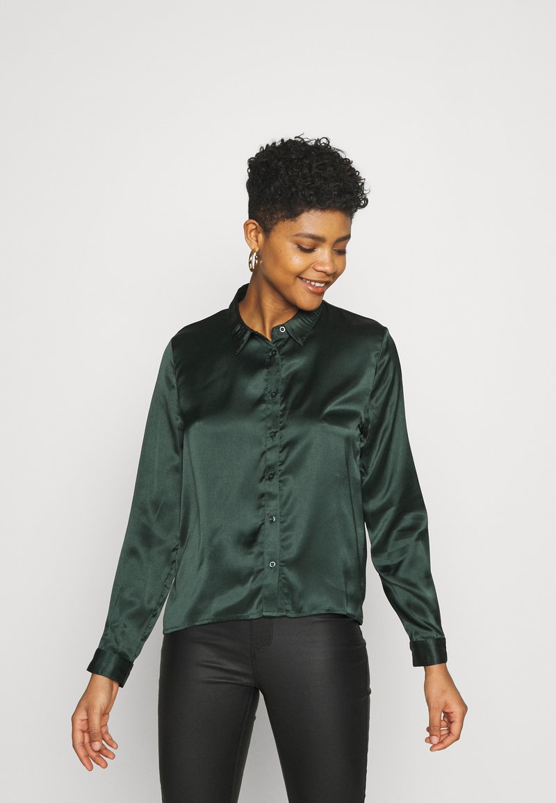 Camicia verde in satin con bottoni, maniche lunghe e colletto classico, caratterizzata da una texture liscia e un leggero lucido.