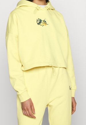 Personne portant un sweat à capuche court jaune pâle avec broderie de citron et feuilles, et un pantalon de survêtement assorti avec taille ajustable à cordon.