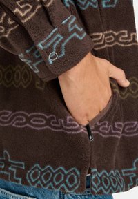 Mano nella tasca di una giacca di pile marrone con motivi geometrici blu, viola e verdi e bottone a pressione sul polsino.