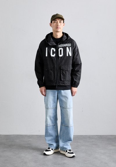 Vyras vilkintis alyvuogių spalvos kepurę, juodą striukę su baltu užrašu "ICON DSQUARED2", šviesiai mėlynas džinsus ir baltai juodus sportbačius, stovi prieš paprastą sieną.