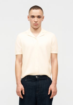 Mladá osoba s krátkymi vlasmi, oblečená v krémovej krátkoročnej košeli na gombíky a tmavomodrých džínsoch, stojaca pred jednoduchým svetlým pozadím.