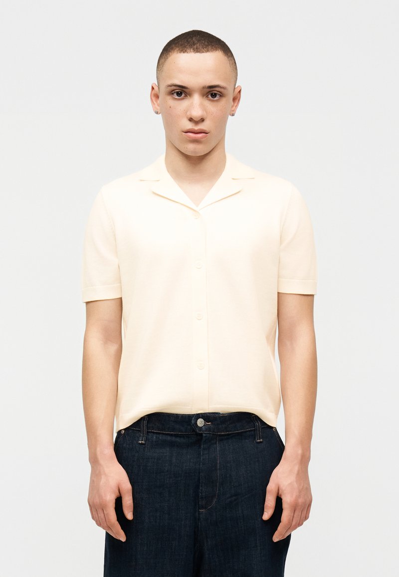 Νέος με κοντά μαλλιά που φορούν ένα κρεμ κοντομάνικο πουκάμισο με κουμπιά και σκούρο μπλε τζιν, στέκεται μπροστά σε απλό φωτεινό φόντο.