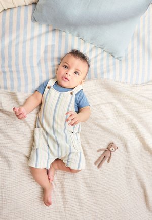 Dieťa v pruhovaných nohaviciach a modrej košeli ležiace na posteli s pruhovanými a béžovými prikrývkami, vedľa malého pleteného plyšového medvedíka.