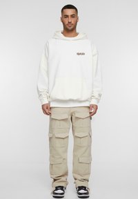 Hvit hettegenser med frontlomme og logo, kombinert med beige cargo-bukser med flere lommer, og svarte og hvite joggesko.