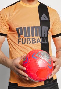 Orange och svart randig Puma fotbollströja med "PUMA FUBBALL"-logotyp, tillsammans med en röd fotboll med blå accenter.