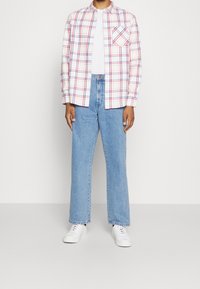 Person som bär en vit skjorta under en uppknäppt vit rutig skjorta med röda och blå linjer, ljusblå jeans och vita sneakers.