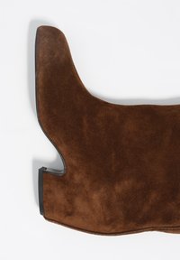 Brązowy zamszowy but o zakrzywionym kształcie, z teksturowaną cholewką i czarną gumową podeszwą. Unikalny wycięty wzór przy kostce.