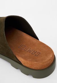 Sandalo in suede verde oliva, con insole texturizzata marrone. Presenta una suola spessa in gomma e un design con punta arrotondata. Il nome del marchio è impresso.