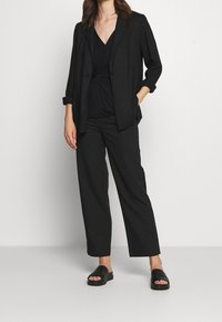 Kvinna klädd i en svart blazer, rynkad svart topp, raka svarta byxor och svarta glidsandaler, stående mot en helt vit bakgrund.