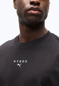 Κοντινό πλάνο ενός ατόμου που φοράει μαύρο Τ-shirt με την επιγραφή "HYROX" και το λογότυπο της Puma τυπωμένα με λευκό χρώμα στο στήθος.
