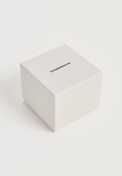 Λευκό κουτί σε σχήμα κύβου με ελαφρώς στρογγυλεμένες άκρες, με τη μάρκα "Completedworks" στο κέντρο της επάνω επιφάνειας.