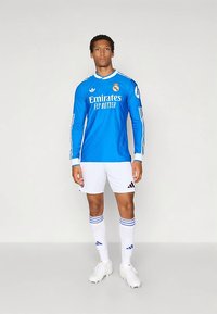 Blaues langärmeliges Fußballtrikot mit weißen Akzenten, Club-Logo und Schriftzug "Emirates Fly Better". Kombiniert mit weißen Shorts und gestreiften Socken.