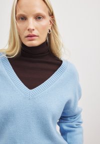 Helles blaues V-Ausschnitt-Pullover aus weichem Strickstoff, über einem dunkelbraunen Rollkragenpullover getragen. Verfügt über einen gerippten Kragen und Ärmelabschlüsse.