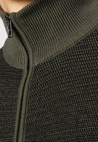 Neulotusta oliivinvihreästä langasta valmistettu vetoketjullinen neulepaita, jossa on tekstuuri neulosta, korkea kaulus ja tyylikäs musta vetoketju edessä.