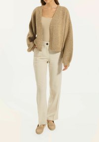 Beige Strickcardigan mit lockerer Passform, V-Ausschnitt und weitem Ärmel; kombiniert mit hellbeigen hochgeschnittenen Hosen und passenden Loafers.