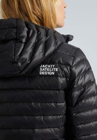 Svart puffjacka med horisontella quiltade mönster, med huva och texten "JACK1T SATELITE DESIGN" i vitt på baksidan.