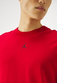 T-shirt de algodão vermelho com um corte clássico de gola redonda e mangas curtas, apresentando um logo Jumpman preto na parte superior do peito. Textura suave.