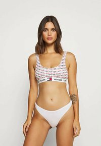 Weißes Bralette mit buntem Druck und Logo-Band, kombiniert mit weißen Bikinihosen. Das Material wirkt glatt mit einem leichten Stretch.