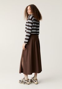 Pruhovaný crop top v tmavej hnedej a bielej farbe, skombinovaný s dlhou hnedou skladanou sukňou s bočným rozparkom a robustnými teniskami.