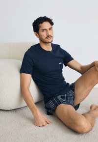 Camiseta de algodão azul-marinho com decote redondo e pequeno logo branco "BOSS", combinada com calções xadrez preto e branco. Fundo suave e texturizado.
