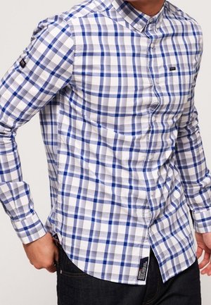 Mężczyzna ubrany w niebiesko-biało-szarą koszulę w kratę z długimi rękawami i ciemne spodnie, ręka spoczywa blisko kieszeni.