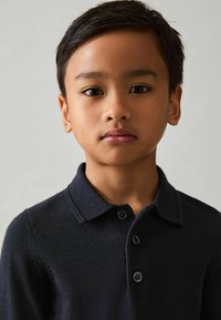 Polo majica tamnoplavih boja izrađena od pletenog materijala s okovratnikom i placket-om na tri dugmeta. Kratki prikaz lica mladog dječaka.