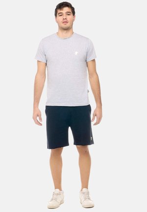 Giovane uomo in piedi con maglietta grigio chiaro, pantaloncini neri e sneakers bianche su sfondo bianco uniforme.