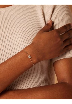 Gouden kettingarmband met een vlinderhanger, versierd met sprankelende details, gedragen om de pols van een persoon met een donkere huid tegen een textuurrijke achtergrond.