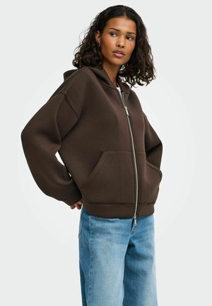 Ung kvinde iført en mørkebrun hættetrøje med lynlås og forlommer og lyseblå jeans, står med hænderne bag ryggen.