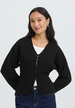 Kvinde med langt mørkt hår iført guldringe-øreringe, sort knaplukket cardigan, hvid top og blå jeans, smilende med hænderne i siden.