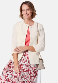 Lichtbeige jas met driekwart mouwen en ronde halslijn, gecombineerd met een koraalkleurige blouse en een geplooide rok met rode en roze bloemenpatronen.