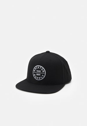 Gorra negra de tela con un parche circular blanco que lleva el texto "Brixton Supply", con forma estructurada y visera plana.