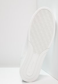 White sneaker with a textured rubber outsole featuring a diamond pattern. Smooth upper material and minimalistic design, shown from the bottom.
