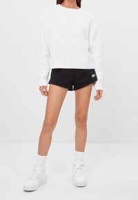 Kvinna i en vit stickad tröja, svarta shorts med en liten grön logotyp, vita ankelsockor och vita högskurna sneakers, stående på ett vitt golv.