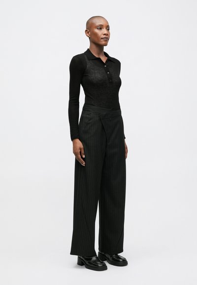 Crna dugačka polo majica s dugim rukavima i dugmadima, u kombinaciji s crnim širokim hlačama s crticama visokog struka i debelim crnim cipelama.