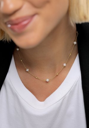 Collier en chaîne dorée présentant des petits accents dorés alternés et cinq perles, posé sur une chemise blanche à col en V.