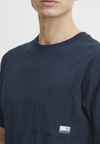Navy blue crew neck t-shirt made of soft fabric featuring a front pocket and a white label with "11 PROJECT" on the hem.
